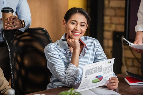 Glückliche afrikanisch-amerikanische Geschäftsfrau mit Infografik lächelt in die Kamera neben Kollegen — Stockfoto