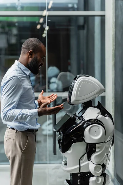 Uomo d'affari afroamericano sorridente gesticolando vicino robot in ufficio — Foto stock