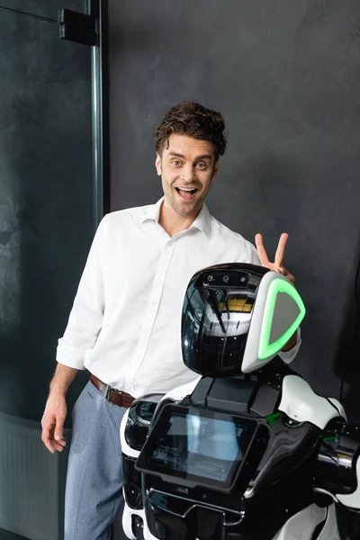 Cheerful businessman looking at camera while showing bull horns sign near robot — Stock Photo