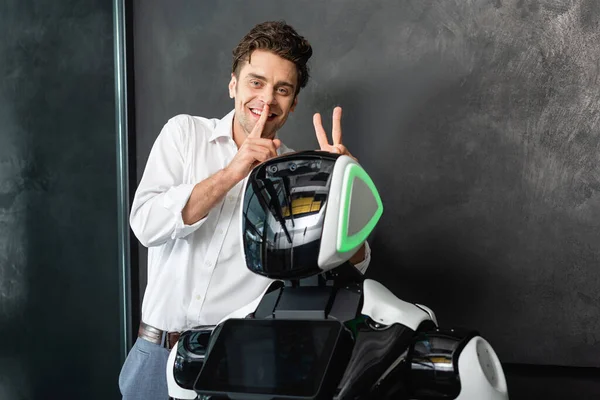 Cheerful businessman showing hush gesture and bull horns sign near robot in office — Stock Photo