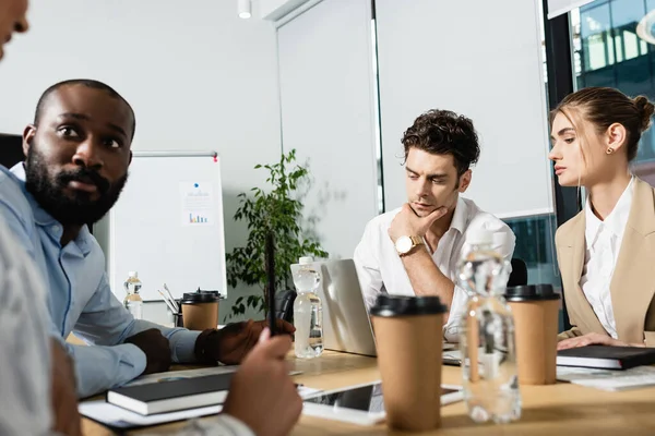 Junge Geschäftsleute denken in der Nähe von Laptop und verschwommenen interrassischen Freunden — Stockfoto