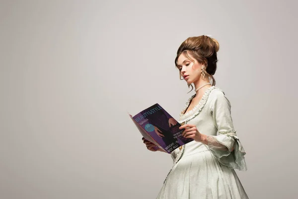 Чарівна жінка в старовинній сукні читає сучасний журнал ізольовано на сірому — стокове фото