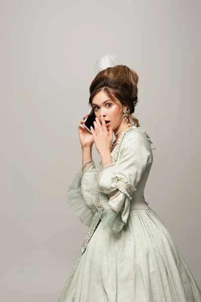 Schockierte Frau im Vintage-Kleid verdeckt Mund beim Telefonieren auf Smartphone isoliert auf grau — Stockfoto