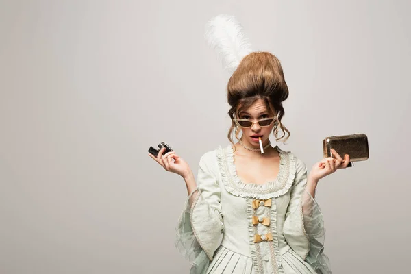 Mujer joven en traje retro sosteniendo bolsa de monedero más ligero y dorado mientras fuma aislado en negro - foto de stock