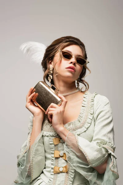 Чувственная женщина в элегантном винтажном наряде и модных солнцезащитных очках позирует с золотой сумкой сцепления, изолированной на сером — стоковое фото