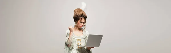 Mujer irritada en vestido vintage mostrando puño cerrado cerca del ordenador portátil aislado en gris, pancarta - foto de stock