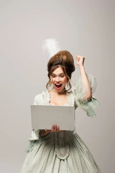Збуджена жінка в ретро-стилі, що показує жест тріумфа, дивлячись на ноутбук ізольовано на сірому — стокове фото