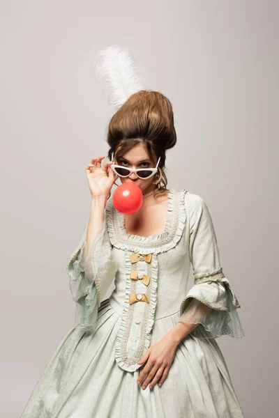 Жінка в стилі ретро в модних сонцезахисних окулярах, що дме жувальна гумка ізольована на сірому — стокове фото