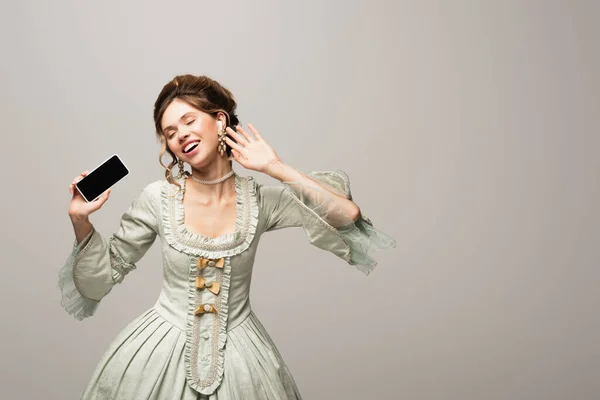 Alegre mujer de estilo vintage escuchando música mientras sostiene el teléfono inteligente con pantalla en blanco aislado en gris - foto de stock