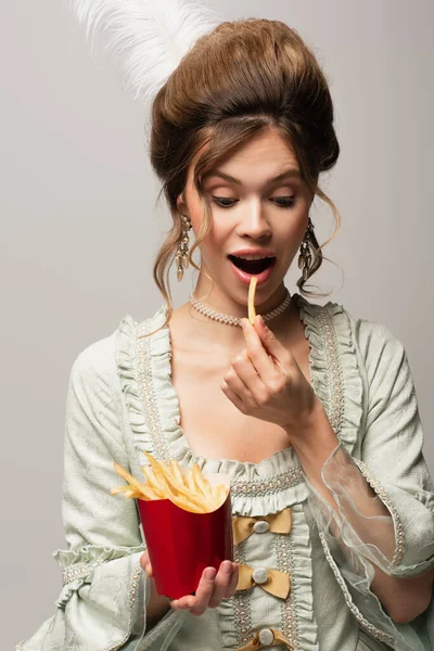 Mujer complacida en elegante atuendo retro comiendo papas fritas aisladas en gris - foto de stock