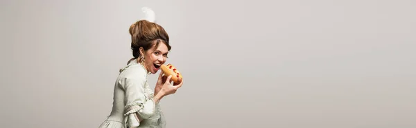 Hungrige, im Retro-Stil gehaltene Frau, die in die Kamera schaut, während sie vereinzelt Hot Dog auf grauem Grund isst, Banner — Stockfoto