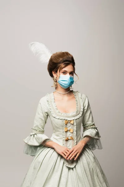 Mujer elegante en vestido vintage y máscara médica mirando a la cámara aislada en gris - foto de stock