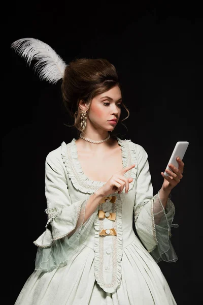Pretty woman in vintage outfit pointing at mobile phone isolated on black — Stock Photo