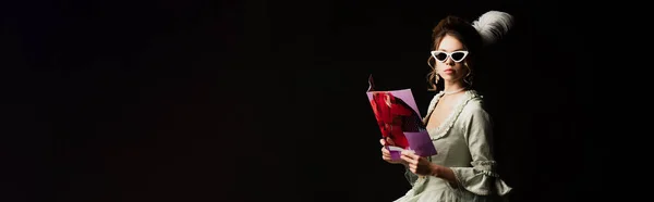 Woman in retro outfit and trendy sunglasses holding modern magazine isolated on black, banner — Stock Photo