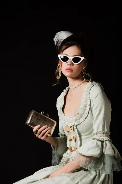 Mujer elegante en vestido vintage y gafas de sol de moda posando con bolso de oro aislado en negro - foto de stock