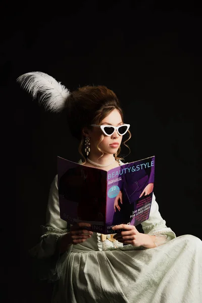 Mujer con estilo en traje vintage y gafas de sol de moda leyendo la belleza moderna y revista de estilo aislado en negro - foto de stock