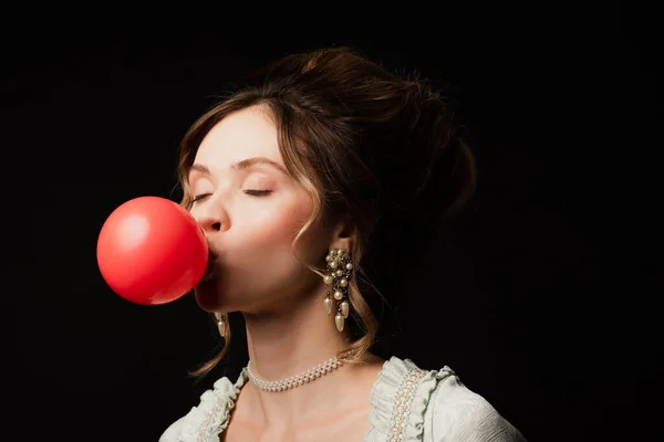 Encantadora mujer de estilo vintage que sopla goma de mascar roja con los ojos cerrados aislados en negro - foto de stock