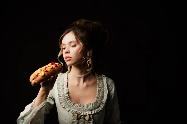 Bonita mujer en elegante vestido retro sosteniendo delicioso perro caliente aislado en negro - foto de stock