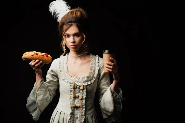 Young woman in elegant retro dress holding coffee to go and hot dog isolated on black — Stock Photo