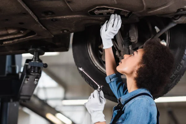 Junger afrikanisch-amerikanischer Mechaniker mit Schraubenschlüssel repariert Boden von angehobenem Auto in Garage — Stockfoto