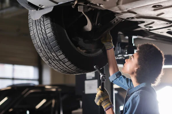 Young african american mechanic with wrench repairing wheel of lifted car in garage — Stock Photo