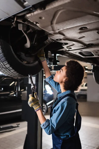 Jovem afro-americano mecânico em macacão segurando chave inglesa e roda de reparação de carro levantado na garagem — Fotografia de Stock