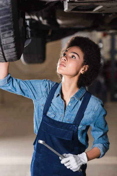 Young african american mechanic with wrench in hand repairing bottom of car in garage — Stock Photo