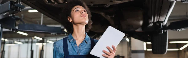 Junger afrikanisch-amerikanischer Mechaniker hält digitales Tablet in der Hand und inspiziert Boden des Autos in der Garage, Banner — Stockfoto