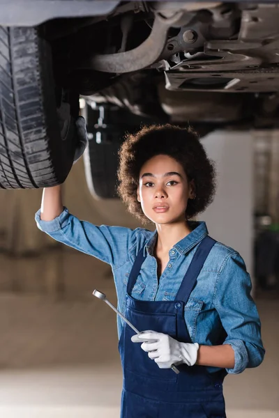 Jovem afro-americano mecânico segurando chave na mão e de pé debaixo do carro na garagem — Fotografia de Stock