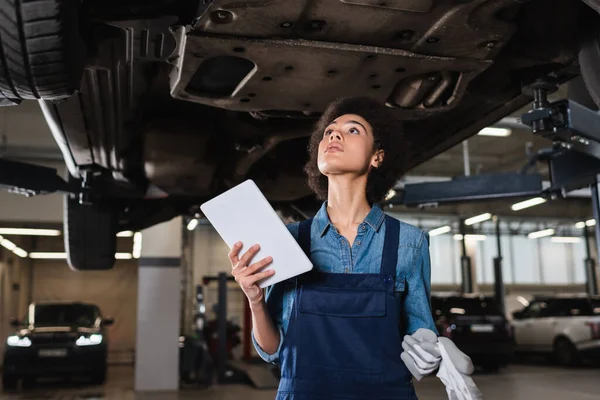 Young african american mechanic holding digital tablet and inspecting bottom of car in garage — Stock Photo