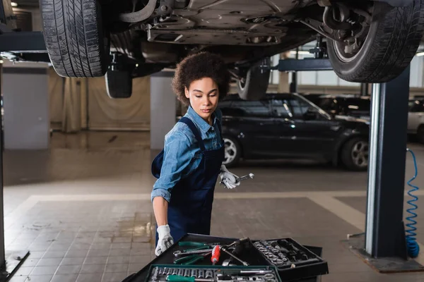 Jovem afro-americano mecânico em macacões reparando carro levantado com instrumentos na garagem — Fotografia de Stock