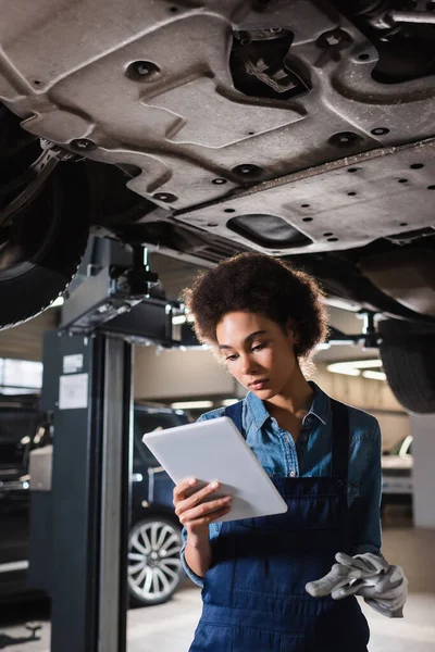 Junger afrikanisch-amerikanischer Mechaniker in Overalls steht unter Auto und hält digitales Tablet in der Garage — Stockfoto