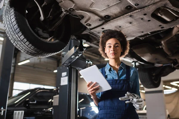 Sonriente joven afroamericano mecánico en overoles de pie debajo del coche y la celebración de tableta digital en el garaje - foto de stock