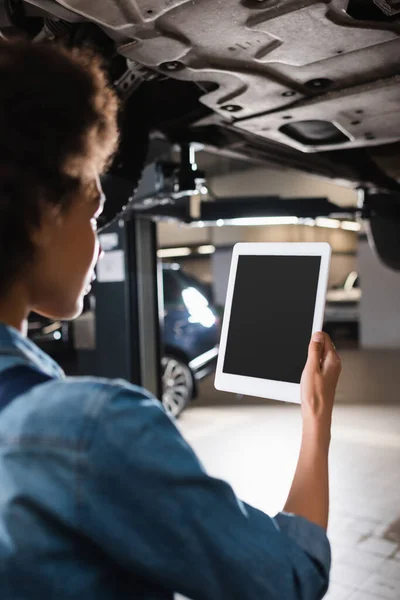 Giovane meccanico afroamericano in piedi sotto l'auto e in possesso di tablet digitale con schermo bianco in garage — Foto stock