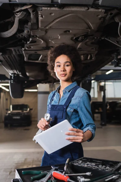 Confiant jeune mécanicien afro-américain debout sous la voiture et tenant tablette numérique dans le garage — Photo de stock