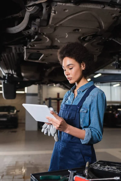 Sérieux jeune mécanicien afro-américain debout sous la voiture et regardant tablette numérique dans le garage — Photo de stock