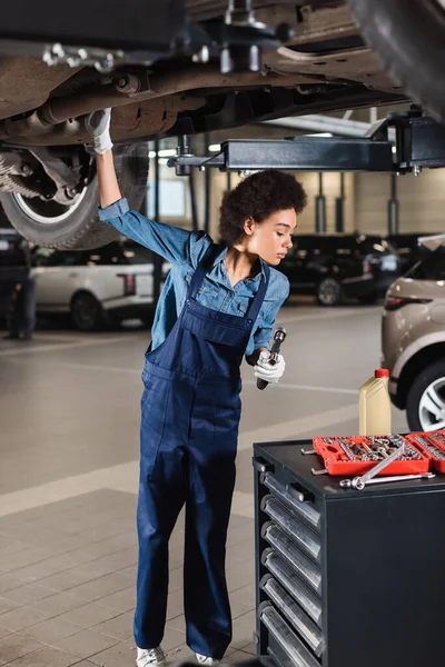 Joven afroamericano mecánico trabajando debajo de coche en garaje - foto de stock