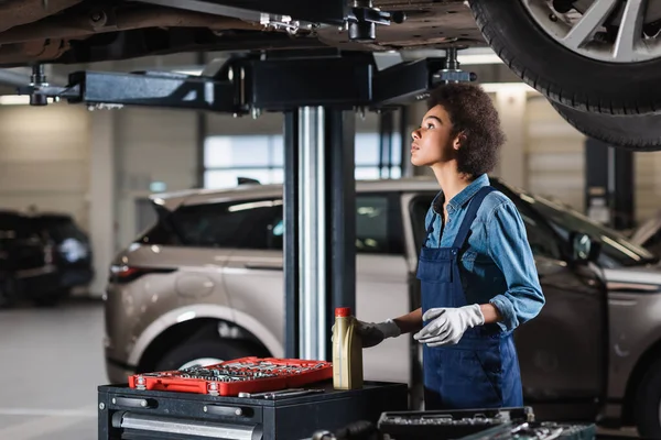 Focused young african american mechanic standing underneath car and holding motor oil bottle in garage — Stock Photo