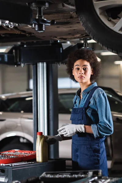 Jovem afro-americano mecânico de pé debaixo do carro, segurando garrafa de óleo do motor e olhando para a câmera na garagem — Fotografia de Stock