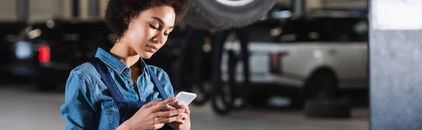 Joven afroamericano mecánico celebración de teléfono celular en las manos en el garaje, pancarta - foto de stock