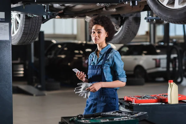 Young african american mechanic standing underneath car and holding cellphone in garage — Stock Photo