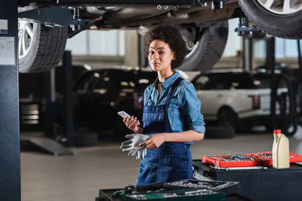 Junger afrikanisch-amerikanischer Mechaniker steht unter Auto und hält Handy in Garage — Stockfoto