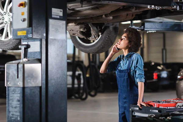 Young african american mechanic standing underneath car and speaking on cellphone in garage — Stock Photo