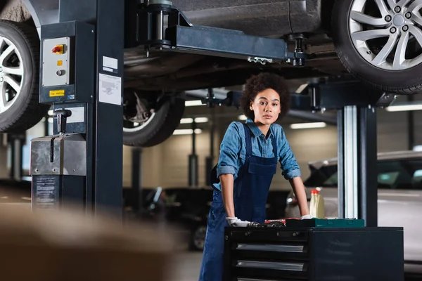 Joven afroamericano mecánico de pie debajo del coche y mirando a la cámara en el garaje — Stock Photo