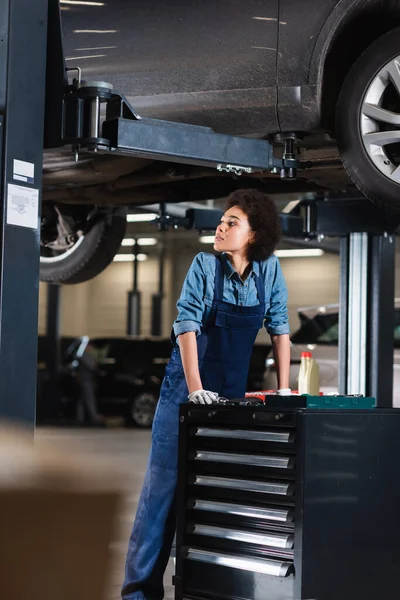 Müder junger afrikanisch-amerikanischer Mechaniker steht unter Auto in Garage — Stockfoto