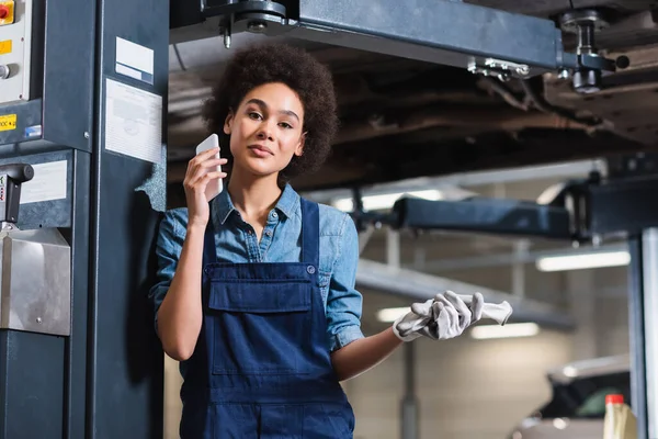 Sonriente joven afroamericano mecánico hablando por celular en garaje - foto de stock