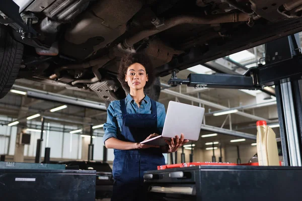 Young african american mechanic standing underneath car, holding laptop and looking at camera in garage — Stock Photo