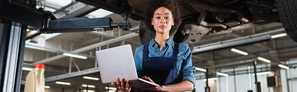 Jovem afro-americano mecânico de pé debaixo do carro, segurando laptop e olhando para a câmera na garagem, banner — Fotografia de Stock