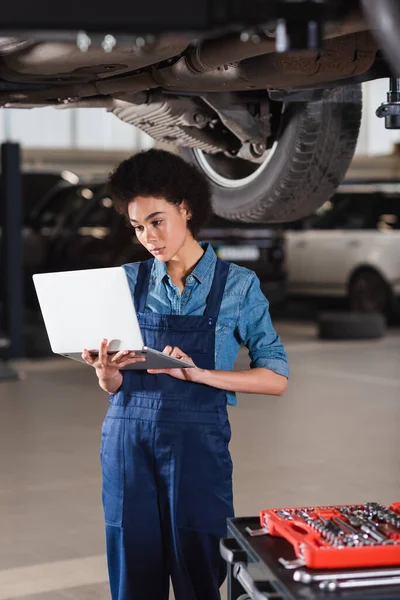 Jovem afro-americano mecânico em pé debaixo do carro e digitando no laptop na garagem — Fotografia de Stock