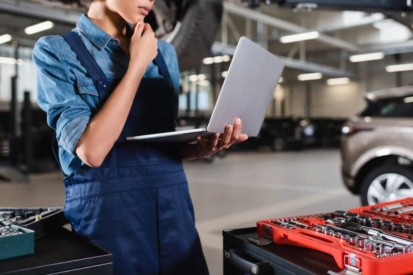 Partial view of young african american mechanic holding hand near face and looking at laptop in garage — Stock Photo